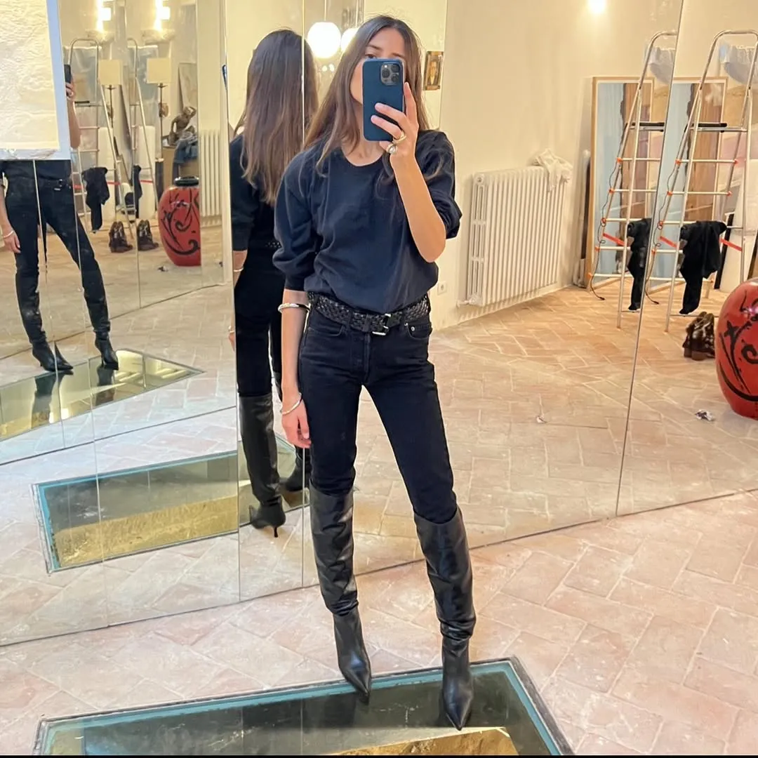 A woman takes a selfie wearing skinny jeans tall boots and a belt