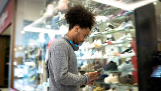 Man looks at phone while standing in front of a store window display of sports shoes