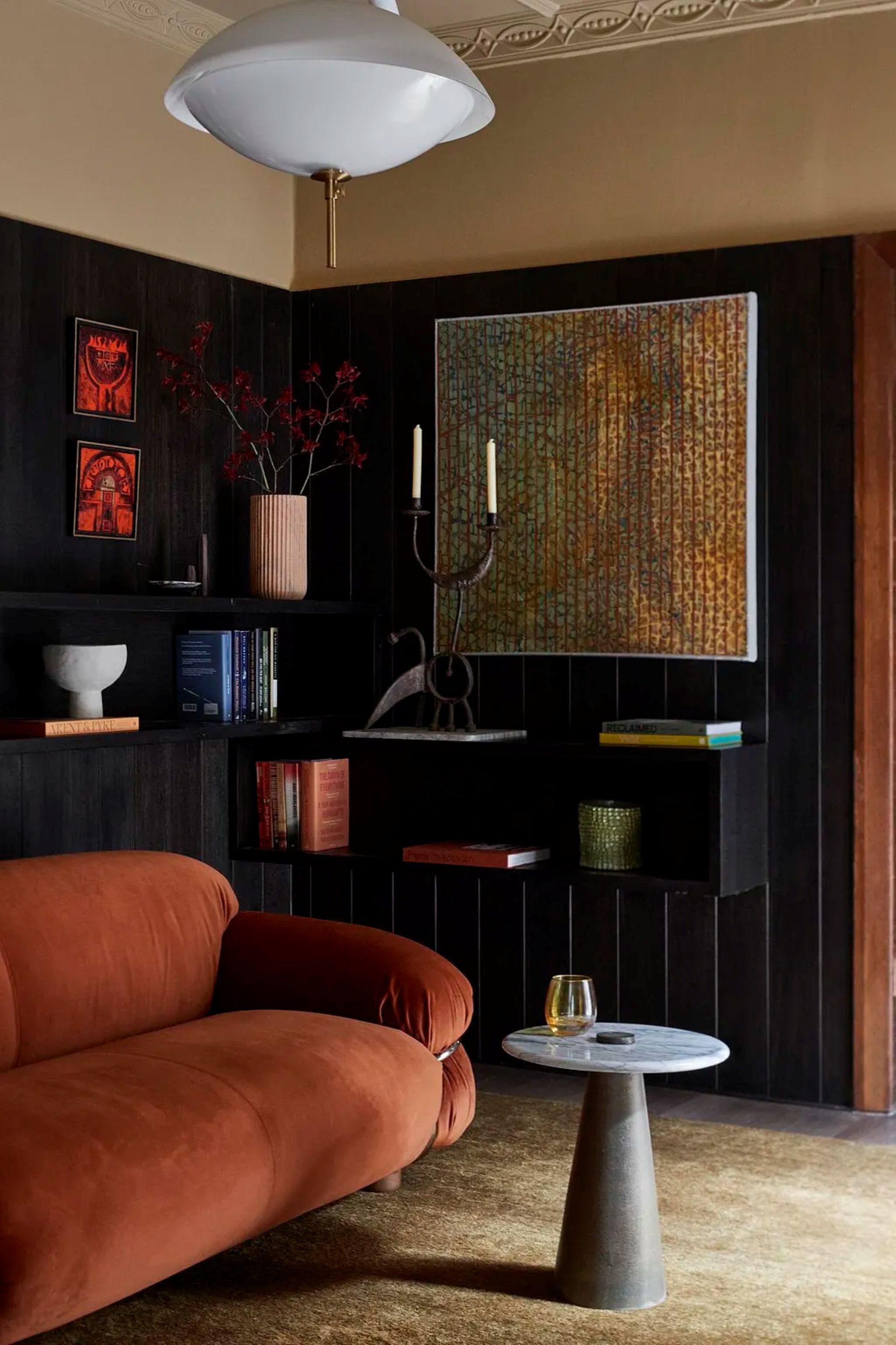 A living room with rust red sofa, black walls, a light tan rug, and a marble small side table. On the wall is various types of wall art including shelves holding flowers, decorative objects, and candles