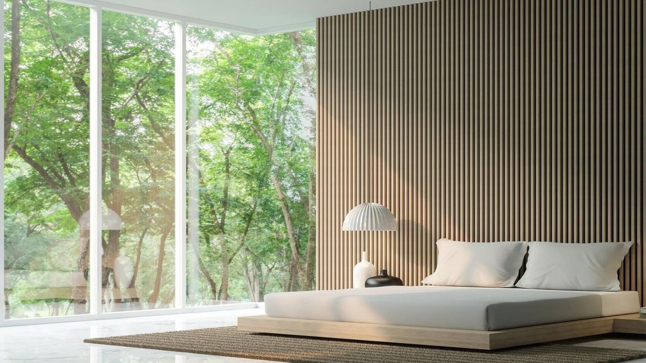 A PlushBeds mattress on a low bed frame against a wood wall and floor-to-ceiling windows.