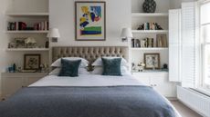 A double bedroom with white bedding with blue accents, and large windows to the right with open white shutters. To either side of the bed are shelves with books, vases, and framed pictures, and above the bed are two white light sconces and a colorful artwork.