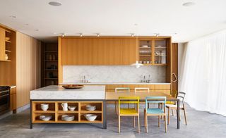 Kitchen in a residential home, with oak wood cabinets/cupboard, white ceiling and grey vinyl flooring. The kithcen has an island with a dining attached to it with 5 colourful chairs