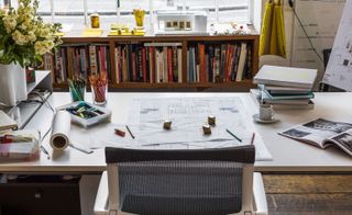 White chair with dark mesh and a white desk with drawings, paperweights, books, pencils, a plant and a small cup with saucer. In the background is a wooden unit filled with books