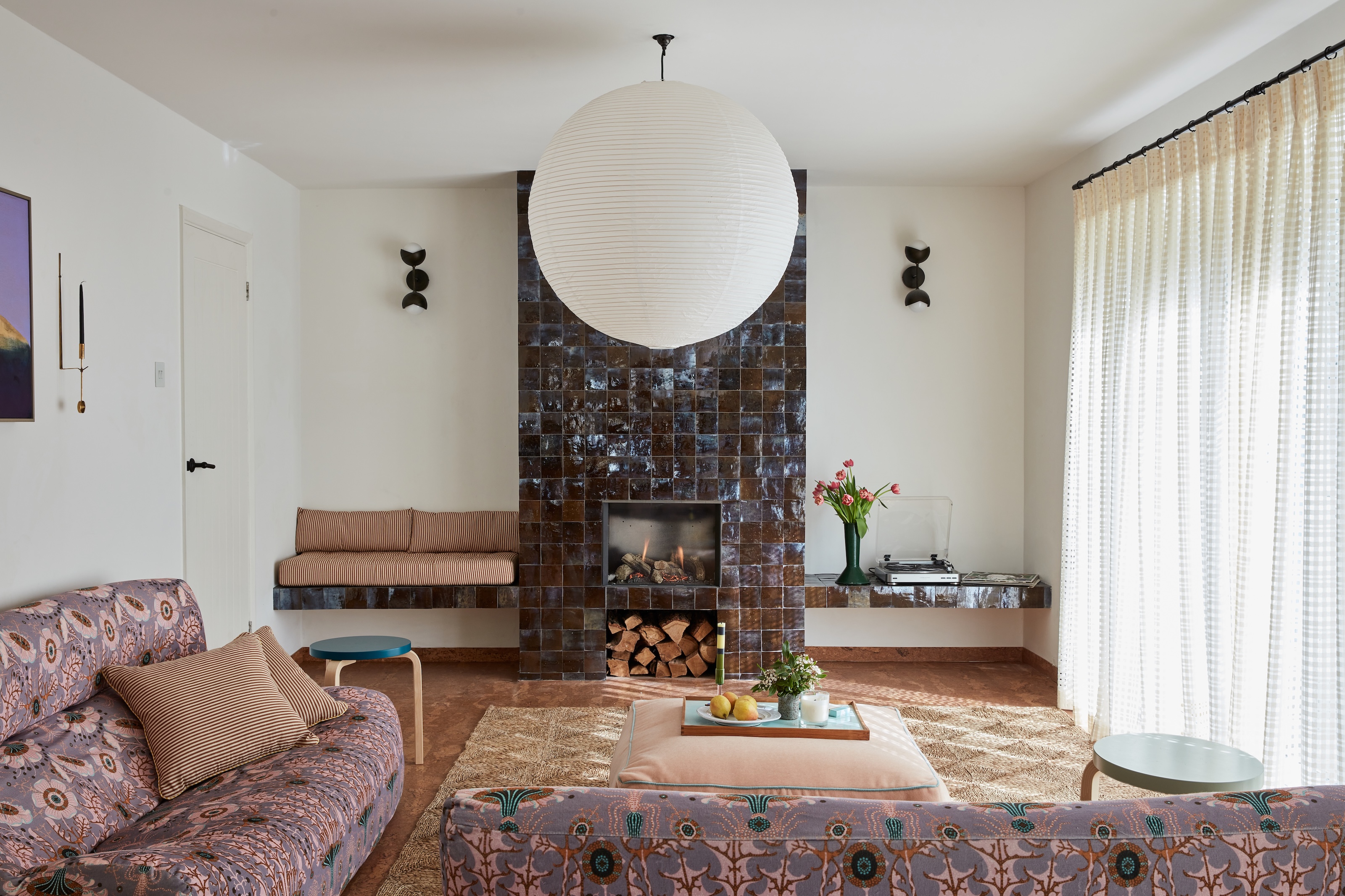 A modern take on a seventies style living room with a chimney breast tiled fully in brown tiles, a bench built in one side and a shelf the other. two sofas in and L-shape just infront with a coffee table and a big paper pendant lamp.