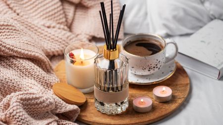 A reed diffuser, candle, and coffee cup on a wooden board on top of a bed with a light pink woven throw and white sheets