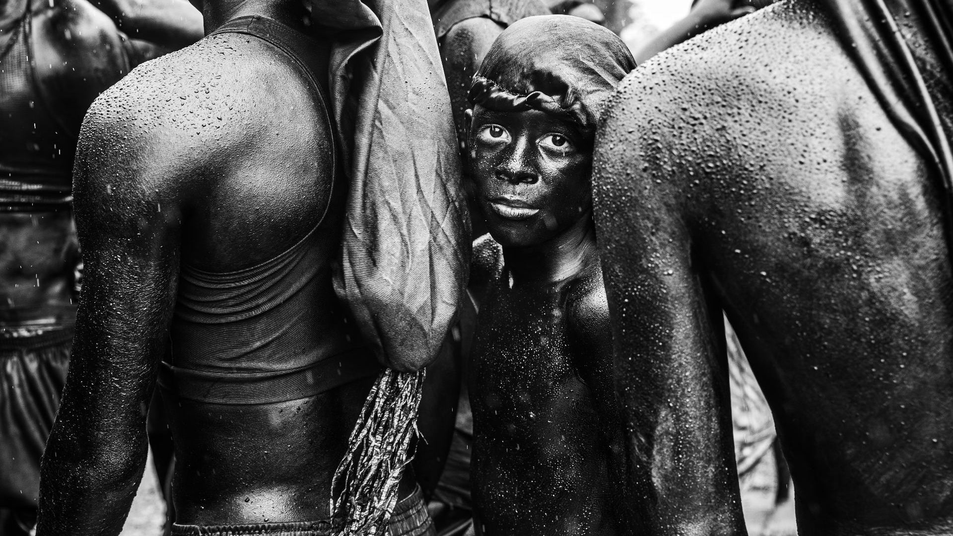Black and white photo of shirtless, wet individuals standing close together, with one young person looking directly at the camera