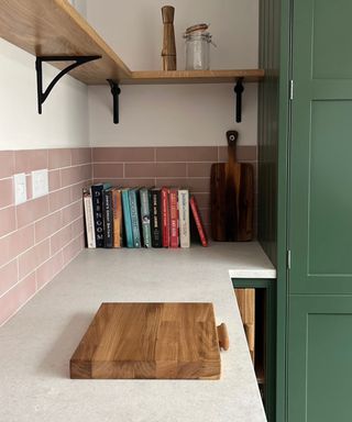 An L-shaped kitchen with wooden open shelves, pink tiles, a white counter with colorful books and wooden chopping boards on it, and a dark green cabinet to the right