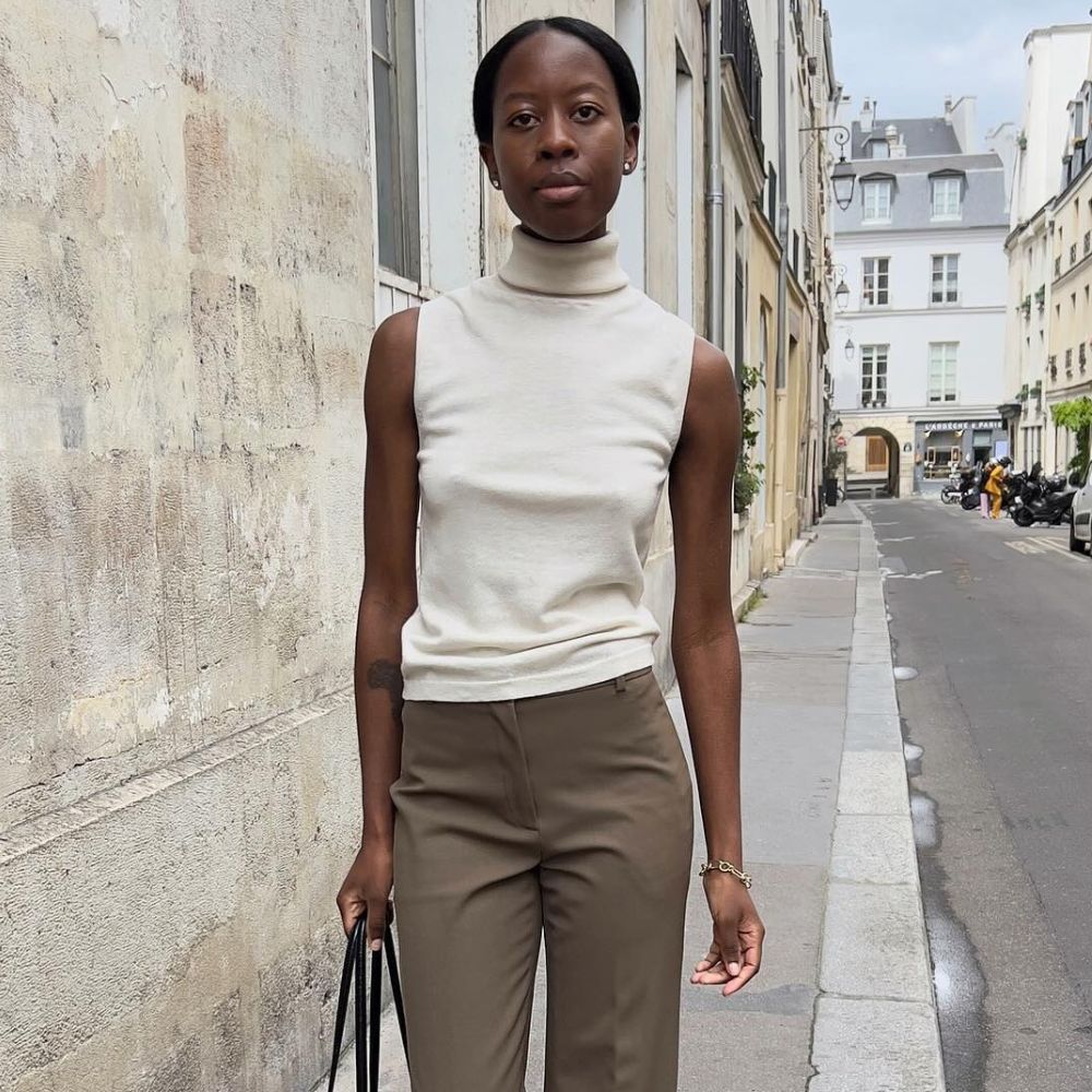 Influencer @sylviemus_ walks outside in Paris wearing low-heel mules in black with an open-toe design with dark khaki trousers, a sleeveless turtleneck kinit and a black bag. 
