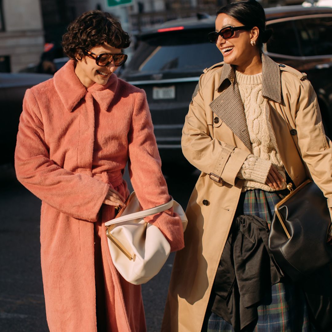 two women smiling at new york fashion week wearing a pink furry coat and a tan trench coat, white cable-knit sweater, and green and blue plaid skirt