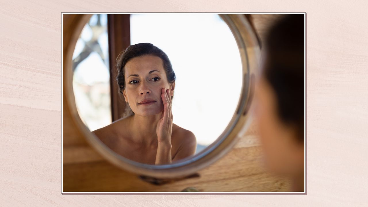 Image of a woman looking at her reflection in a circular mirror hanging on a wooden wall, in a white frame and set against a pale dusky pink watercolour-style background