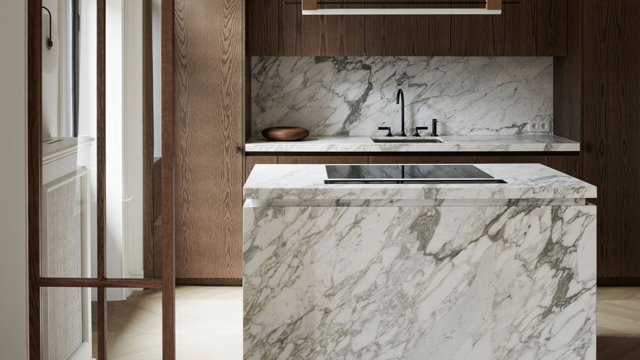 Close up of a square marble kitchen island. Surrounding cabinetry is in dark oak, and the backsplash is in a matching marble 