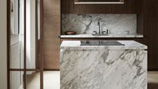 Close up of a square marble kitchen island. Surrounding cabinetry is in dark oak, and the backsplash is in a matching marble 