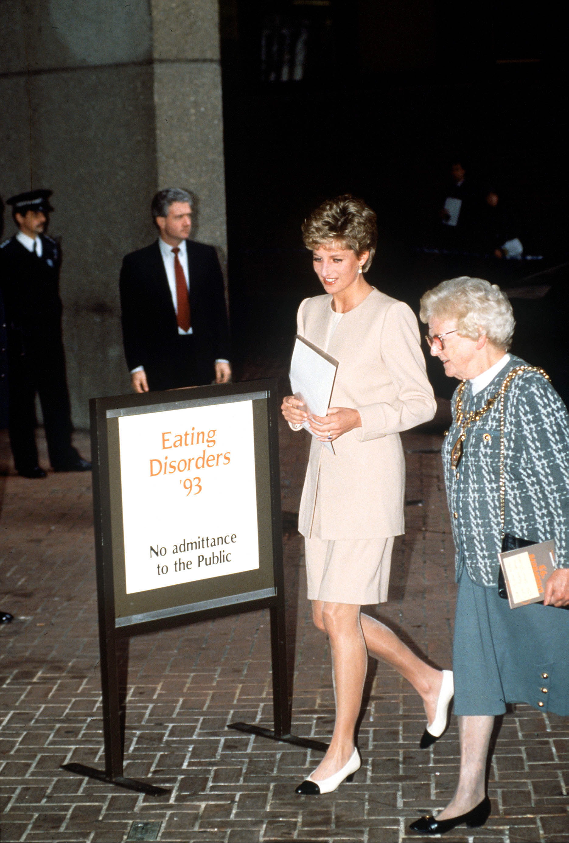 Princess Diana wearing a skirt suit walking past a sign that says Eating Disorders '93
