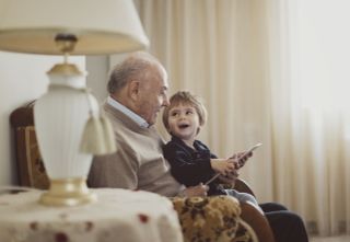 A grandad sat on the sofa with his grandchild who is looking up at his grandpa and holding an electronic tablet.