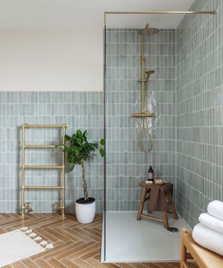 A modern small bathroom with light blue tiles, a clear glass shower with a wooden stool and gold shower head, a gold shower rail with a tall plant next to it, and a wooden table with three folded white towels