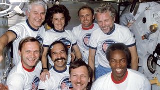 A group of astronauts, smiling and wearing matching "Spacelab" mission shirts, pose together inside the spacecraft. The crew appears happy and relaxed, capturing a moment of camaraderie during their mission.