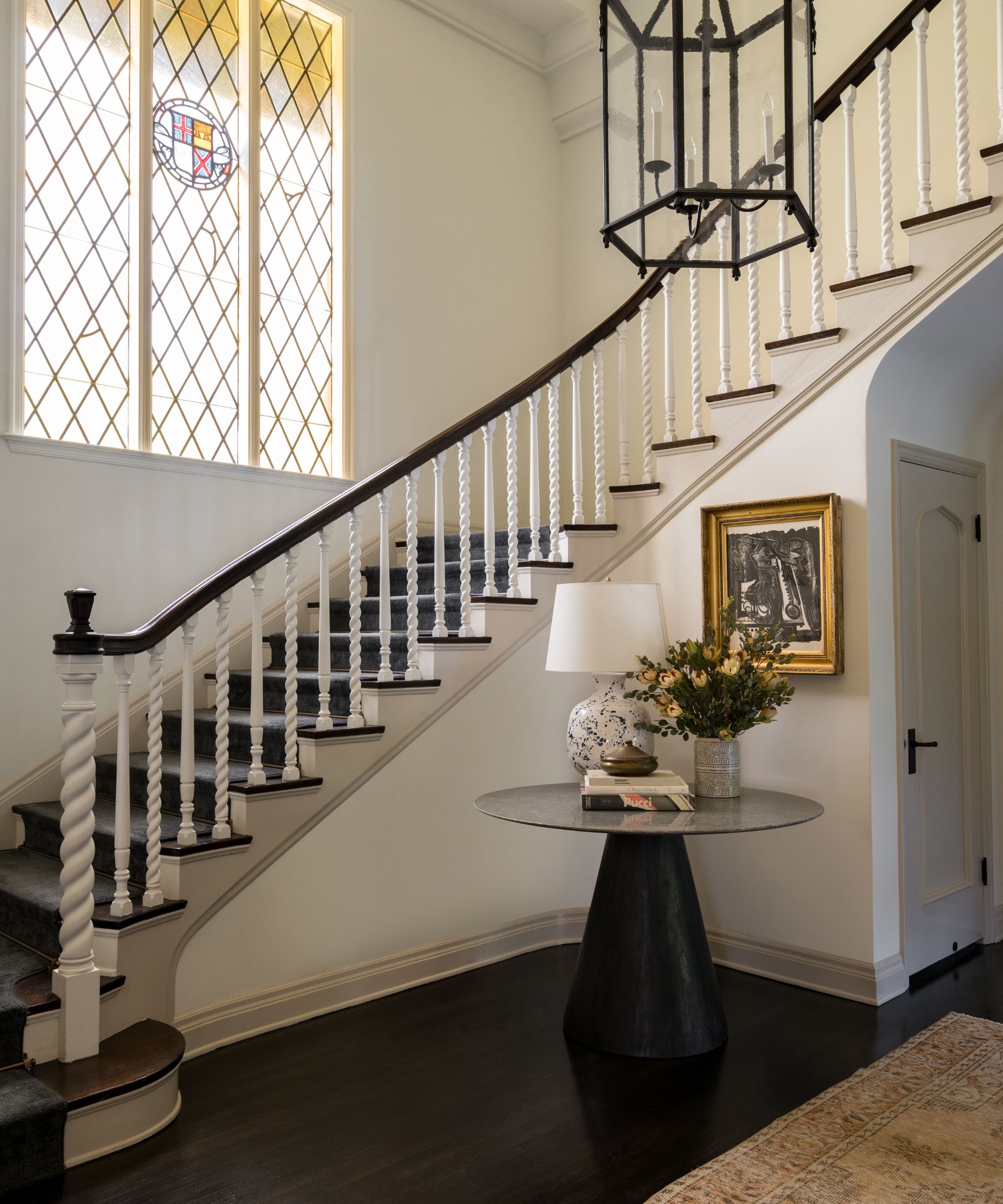 Grand entryway with a large stained glass window and sweeping spindle staircase, a glass lantern light, a round table holding a lamp and flowers, and a painting above it, with a vintage rug below.