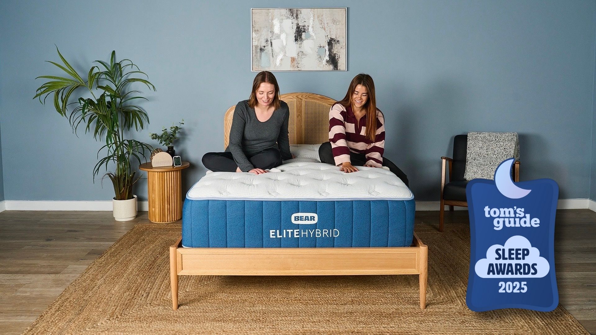 Two of our mattress testers sitting on the navy and white Bear Elite Hybrid mattress on a wooden bedframe with a rattan headboard in a sleep studio with a blue wall, wooden bedside table, a plant and black chair