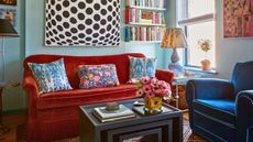 Small and cozy eclectic colorful living room with alcove shelving filled with books, orange and blue velvet sofas, and large black and white geometric artwork on the wall. There is a bright window and a coffee table with books and a vase of flowers