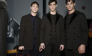 Three male models wearing looks from Giorgio Armani's collection. Two models are wearing crew neck tops, black suits and waistcoats. The third model is holding a bag and is wearing a blue and black collarless shirt, blue trousers and a black coat