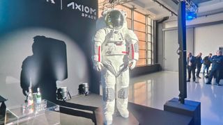 photo of a white spacesuit standing on stage at a conference, with a handful of onlookers in the background