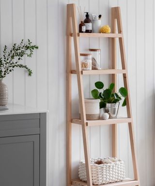 A white bathroom with a wooden ladder shelf with decor on it next to a gray cupboard