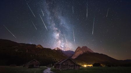 a mountain peak in the foreground in darkness. behind are shooting stars streaking toward the horizon. the milky way is glowing in the background in the vertical middle of the image