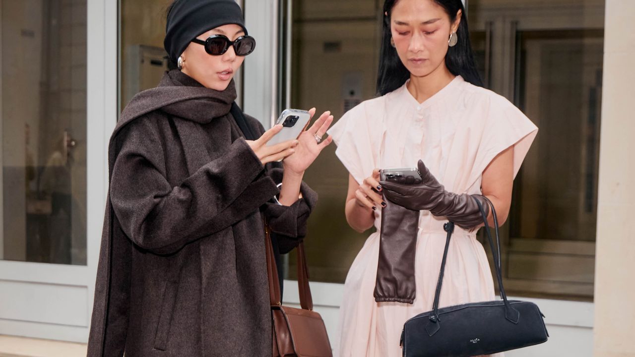 a close street style photo of two women standing outside on the sidewalk scrolling their phones in search of the best ssense sale finds 