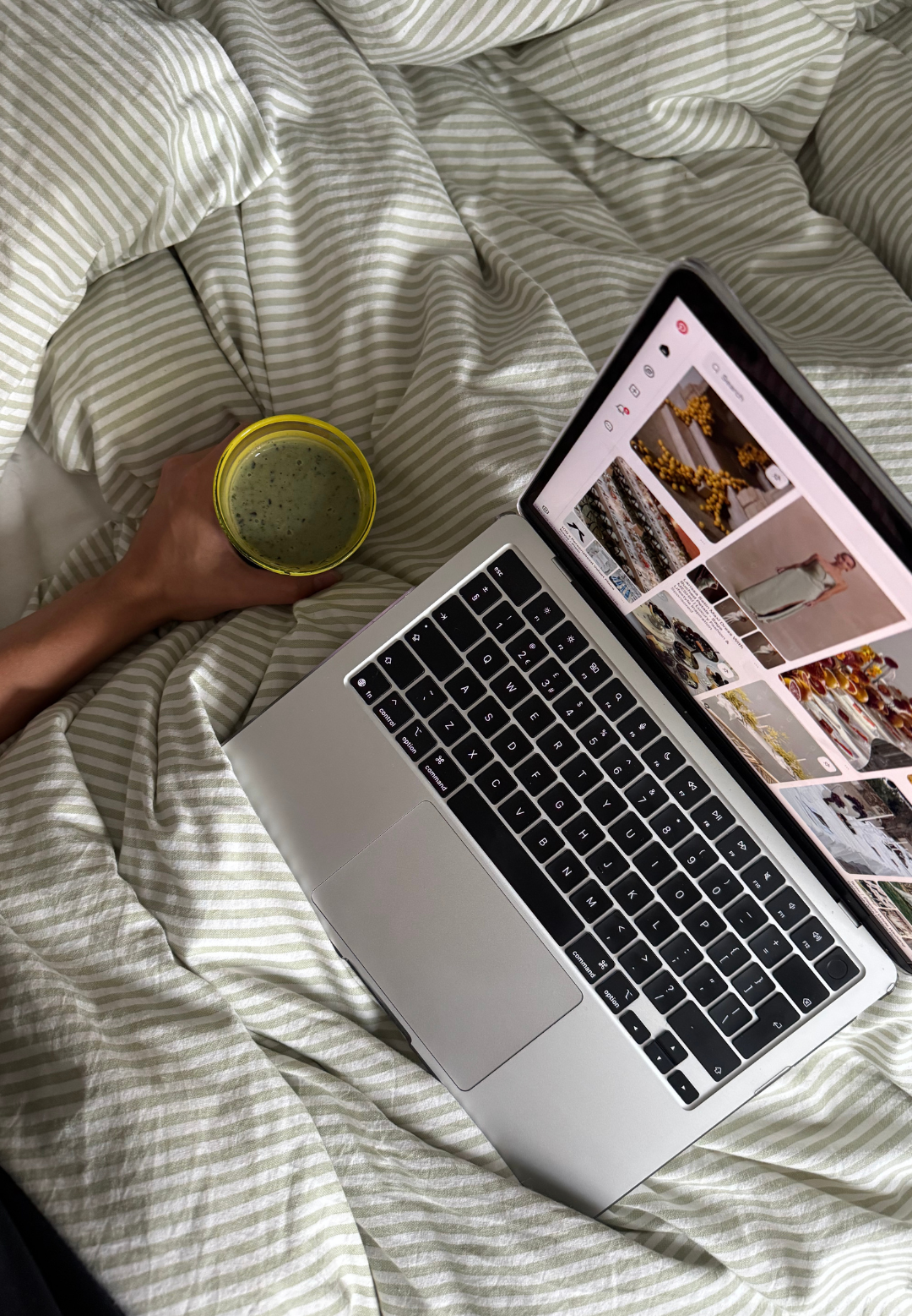 Hand holding a green smoothie on striped green bedding with a laptop