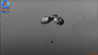 a space capsule floats through the sky beneath three parachutes.