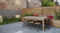 A patio with a wooden slatted fence, a wooden table and chairs, and a border of green plants