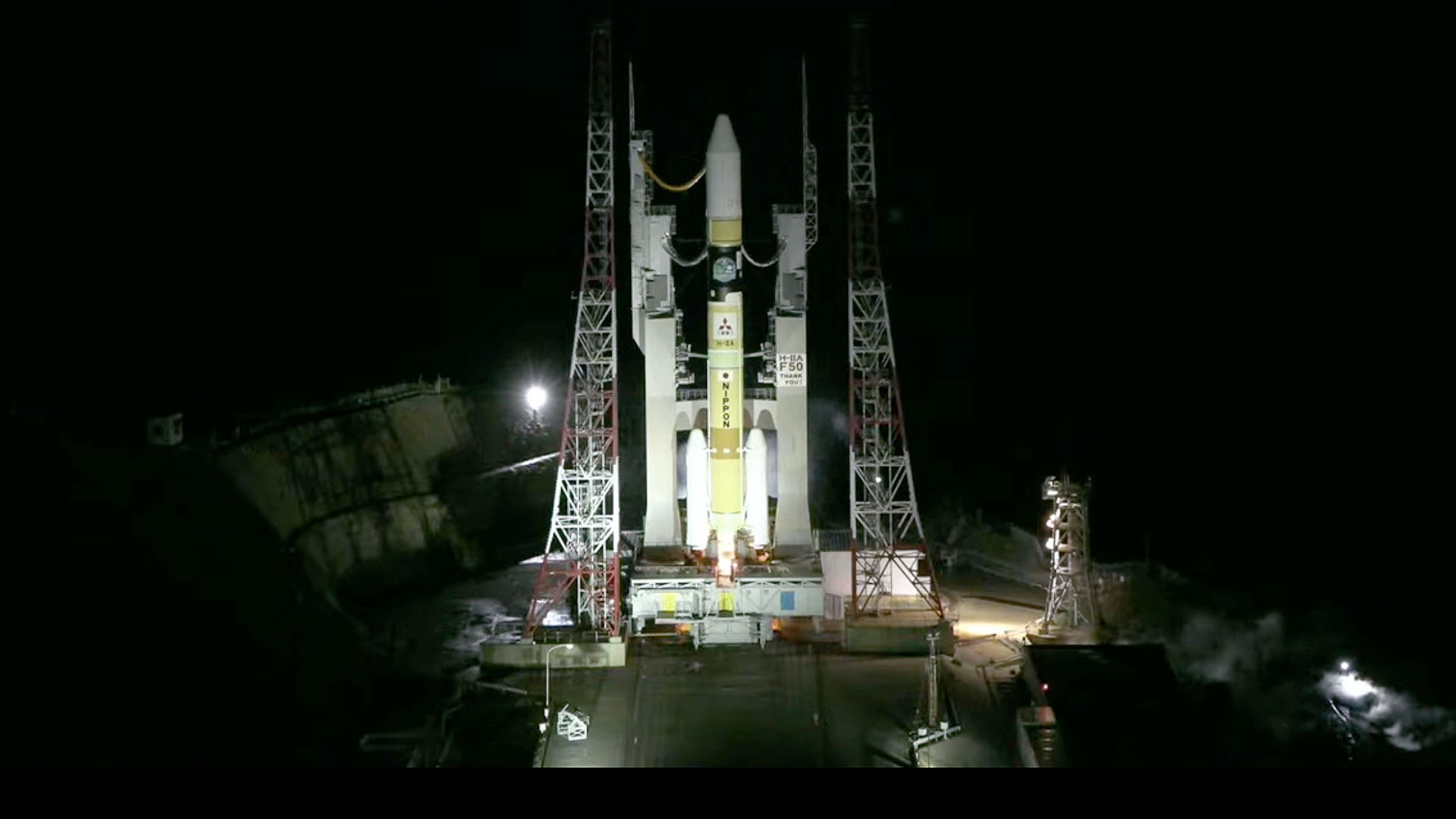 an orange and white rocket stands poised on its launch pad under a black night sky.