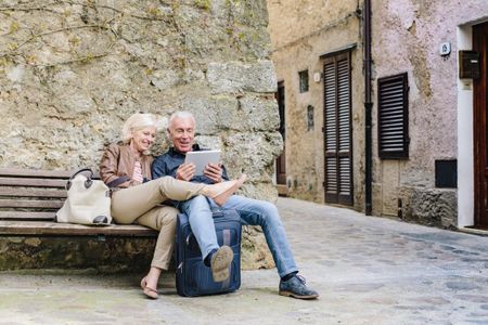 A mature couple sit on a bench looking at a digital notepad.