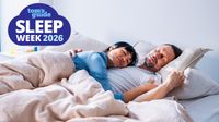 A woman dressed in a blue night shirt sleeps peacefully on her partner's chest, as he sleeps with his head on a stone colored bed pillow. In the top left hand corner is a blue Tom's Guide Sleep Awareness Week 2026 logo.