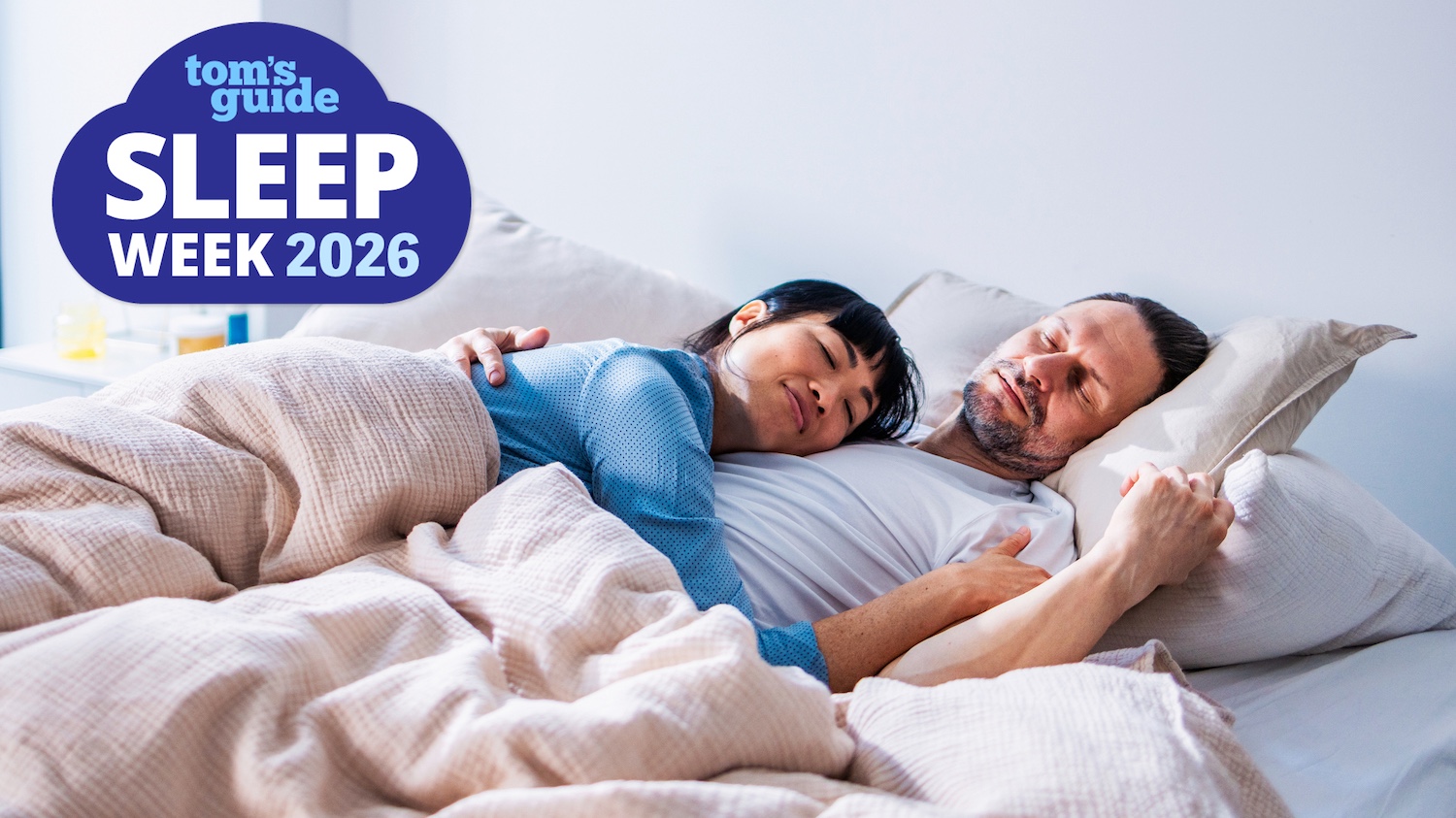 A woman dressed in a blue night shirt sleeps peacefully on her partner's chest, as he sleeps with his head on a stone colored bed pillow. In the top left hand corner is a blue Tom's Guide Sleep Awareness Week 2026 logo.