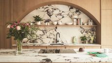 A white marble kitchen island counter and alcove backsplash with dark gray-brown veining paired with natural wood accents with decor piece on the shelves and flowers in a vase on the island