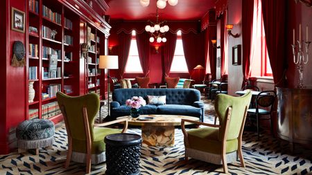 A red painted living room with red curtains on high windows. There is a zebra print carpet that extends over the whole room. There are multiple sitting ares with a blue sofa, green fringe accent chairs, and floor to ceiling bookshelves. 