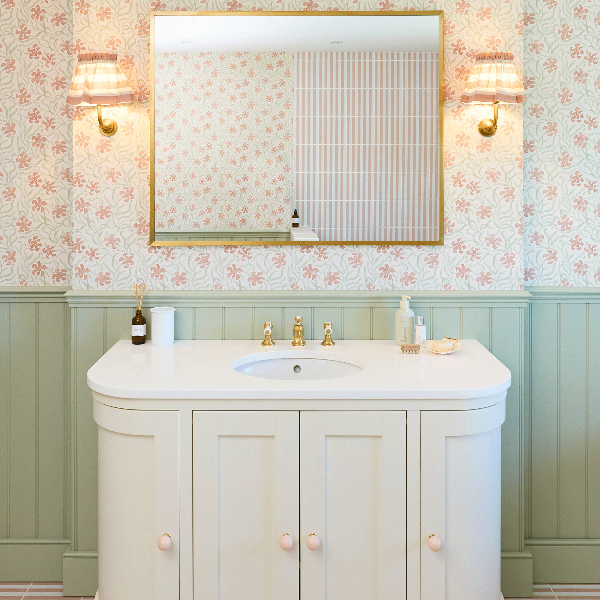 Bathroom with green wall panelling, floral wall paper, pink and white stripe tiles in shower enclosure, stripe wall lights with a gold mirror over a white vanity