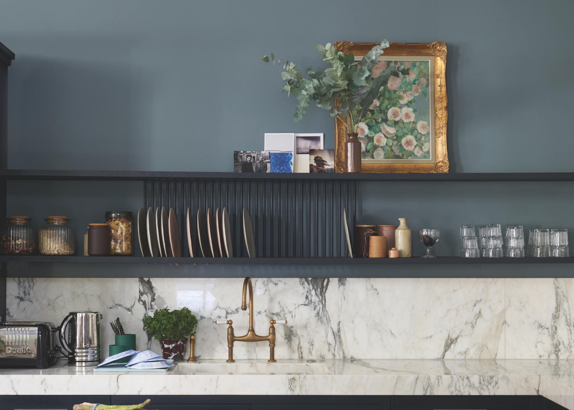 modern kitchen with blue walls and marble counters, there&#039;s a dish rack above the brass sink and open shelving