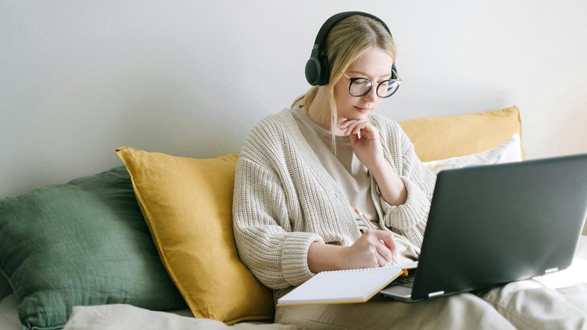 A woman in bed wearing headphones and working on a laptop while taking notes