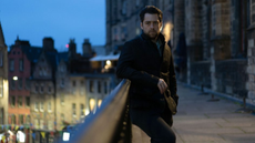 Richard Rankin as Detective John Rebus leans on a railing in Edinburgh at nightfall.