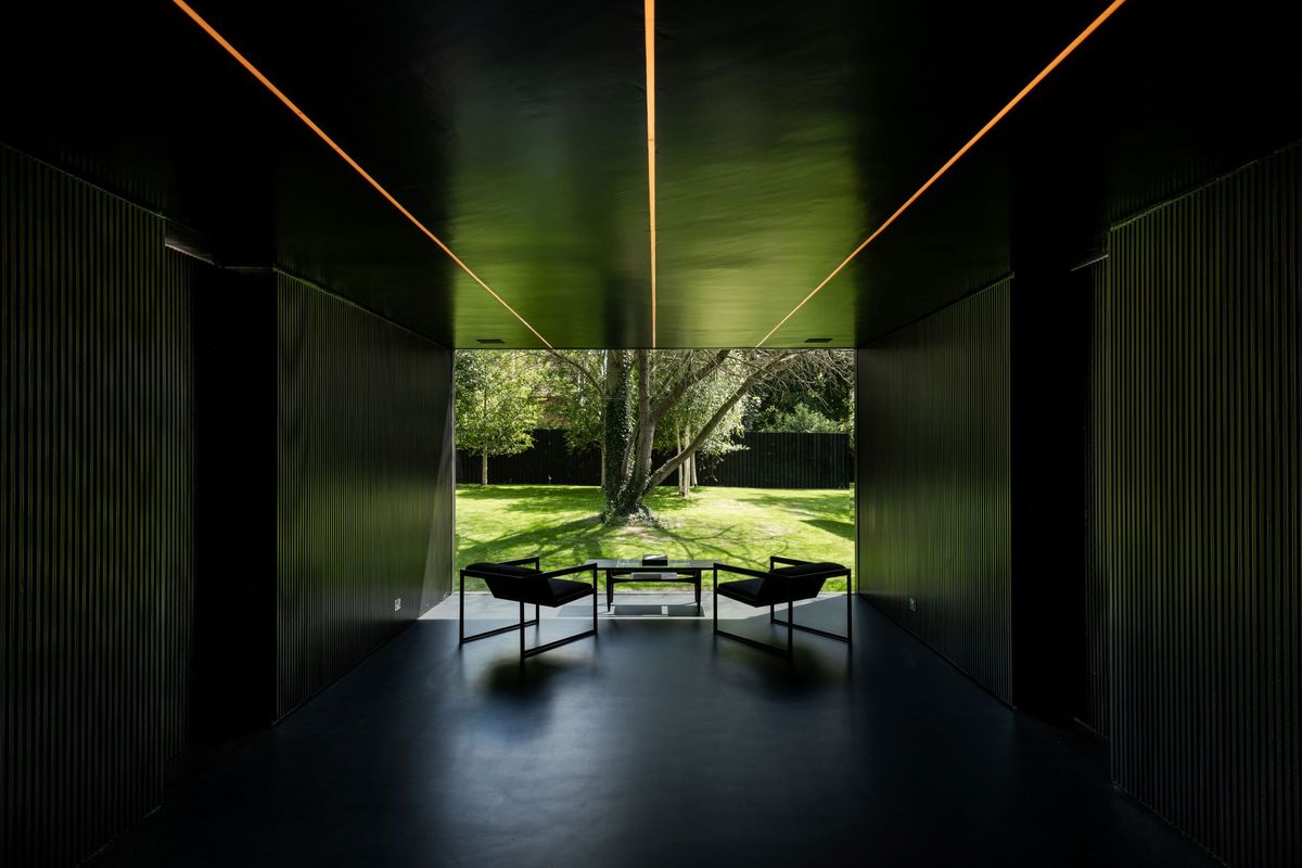 A black room with three strips of light on the ceiling pointing to a tree in the garden which can be seen through the window