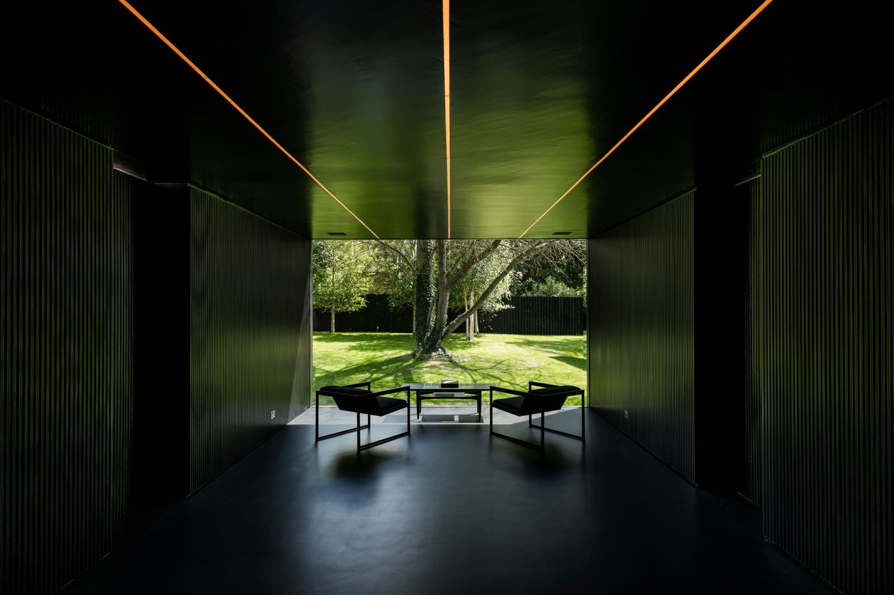 A black room with three strips of light on the ceiling pointing to a tree in the garden which can be seen through the window