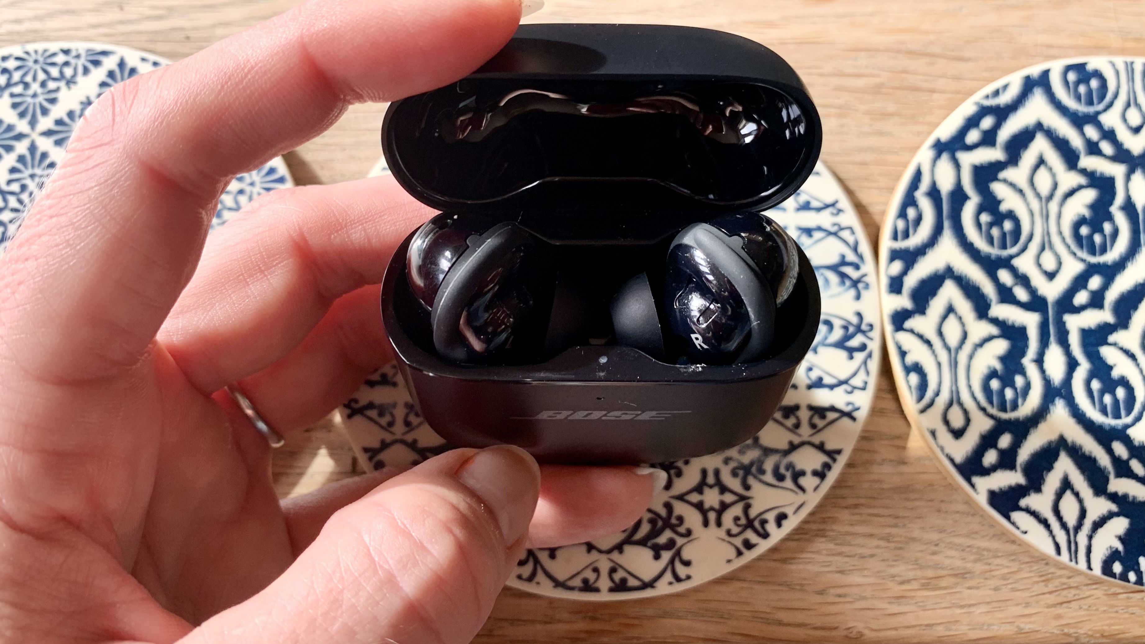 Bose QuietComfort Ultra Earbuds in case, open, held in a hand, on a coffee table