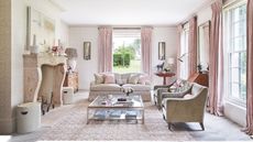 A pink living room with pink curtains, a pink patterned rug, glass coffee table, neutral couches and a large stone fireplace. The walls are white and a green garden can be seen through the windows along the walls. 