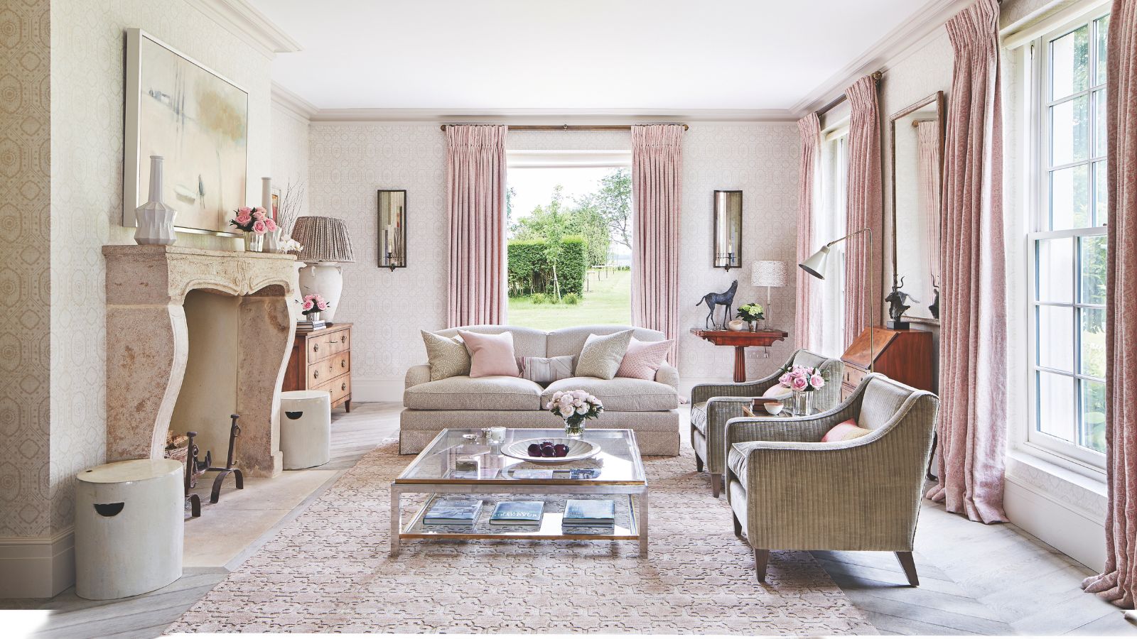 A pink living room with pink curtains, a pink patterned rug, glass coffee table, neutral couches and a large stone fireplace. The walls are white and a green garden can be seen through the windows along the walls. 