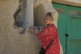 Jean (Sally Lindsay) stands with her hands leaning against a stone wall, next to set of green doors which appear to lead to some sort of storage room or workshop