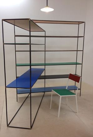 Interior room, white walls, neutral floor. black framed desk unit, blue, black, green and red shelving, white framed desk chair with green seat and red back, black frame ceiling light, white shade, lit bulb