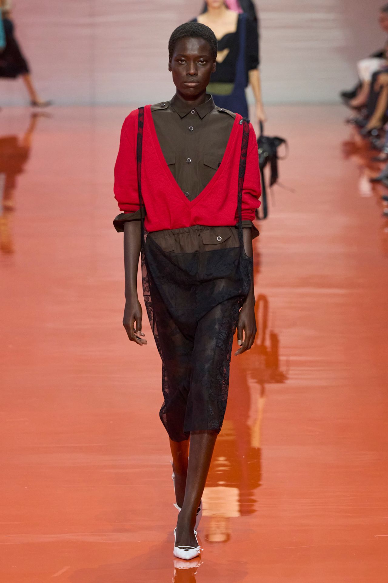 A Prada model wearing a brown button-down shirt layered underneath a bright red V-neck sweater, dark brown shorts, a sheer black slip skirt, and white pointed pumps.