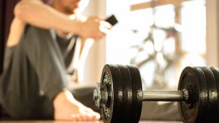 Dumbbell on floor, man in background sitting on the floor looking at phone